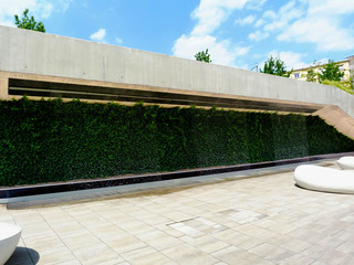 Public street with display of park and decorative water curtain and green ivy. grey concrete wall frame and white stone seats on stone sidewalk pavement. light blue sky and white clouds. urban scene