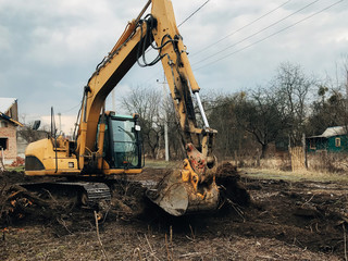 Excavator uprooting trees on land in countryside. Bulldozer clearing land from old trees, roots and branches with dirt and trash. Backhoe machinery. Yard work