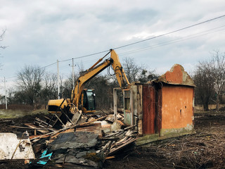 Excavator destroying brick house on land in countryside. Bulldozer clearing land from old bricks and concrete from walls with dirt and trash. Backhoe machinery ruining house