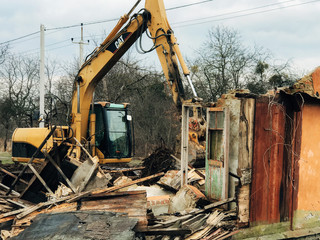 Excavator destroying brick house on land in countryside. Bulldozer clearing land from old bricks and concrete from walls with dirt and trash. Backhoe machinery ruining house