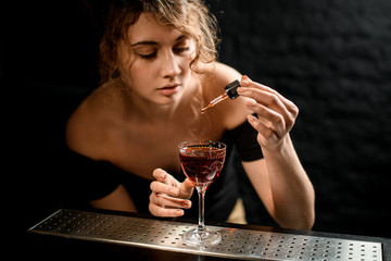 Young attractive female barman adds ingredients to cocktail and looks at it