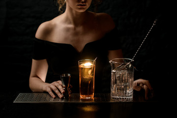 glass with bright alcoholic drink, mixing cup with spoon and metal jigger stand on bar counter.