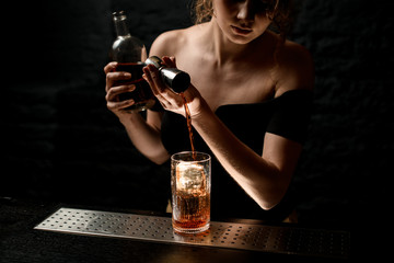 professional female bartender pours amber drink from jigger into glass.