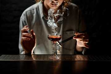 young woman bartender holds tweezers with smoking stick near glass