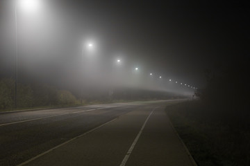Empty road and sidewalk under white street lights in night. Dark time. Foggy air. Poor visibility.