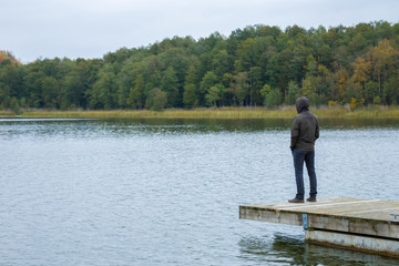 One young man standing alone on edge of wooden footbridge and staring at lake. Hooded guy. Peaceful atmosphere in nature. Enjoying fresh air in cold day. Back view.