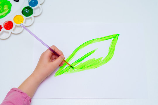 Top view of child is drawing green leaf by watercolors on white paper. Earth day and environmental protection concept.