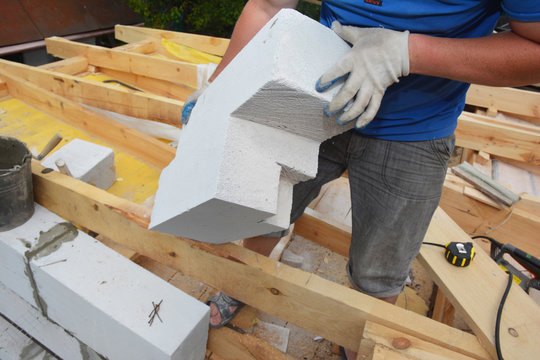 A building contractor installing an autoclaved aerated concrete block of irregular form, cut by a handsaw to lay it on the edge of a brick wall of the house construction where roof trusses installed.
