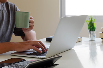 An Asian man uses a laptop at home while sitting at a wooden table. Man hands typing on a notebook keyboard and holding a coffee mug. The concept of young people working at mobile devices.