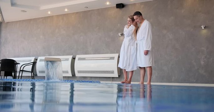 romantic couple in white robes hugs standing on swimming pool edge and playing touching water by feet slow motion