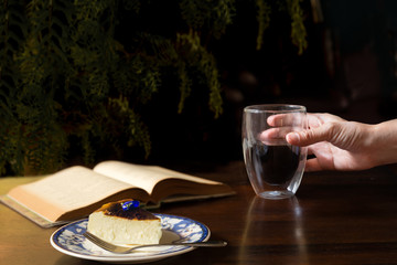 Female hands hold cup of coffee with piece of cake and open book on table.