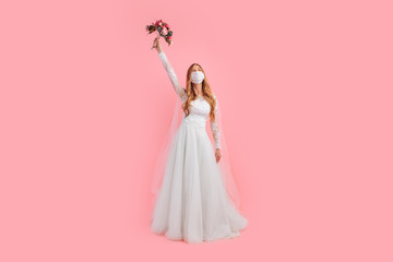 A girl-bride with a bouquet of flowers in a wedding dress and a medical protective mask on her face, on a pink background. Wedding, quarantine, coronavirus