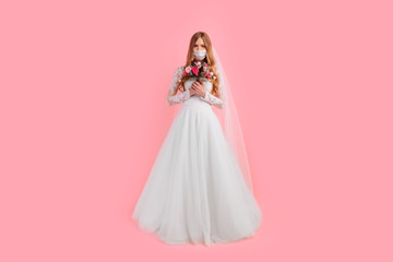 A girl-bride with a bouquet of flowers in a wedding dress and a medical protective mask on her face, on a pink background. Wedding, quarantine, coronavirus