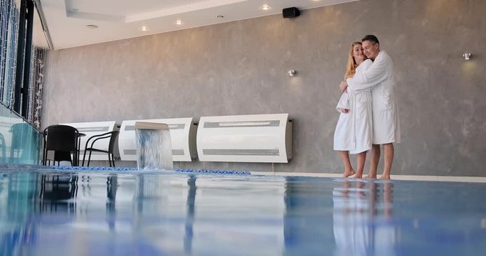 man in white robe approaches young blonde woman and hugs standing near blue swimming pool clear water slow motion
