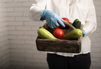 Donation food box in woman's hands in medical gloves. Zucchini, tomatoes and cucumbers in a box, humanitarian aid