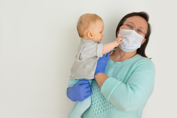 Positive mature woman wearing medical mask and gloves holding baby in arms isolated against white background. Epidemic or virus guidance concept