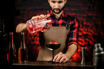 young male barman carefully pours cocktail from mixing cup into black glass.
