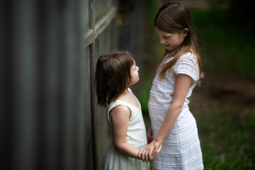 Fototapeta premium Tender young girls children hold hands and laugh near a wooden fence, intimacy and openness, trust between sisters. The concept of the relationship of siblings and childhood., Sincere emotions.