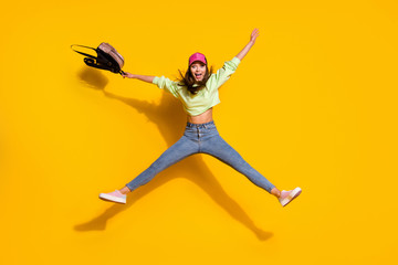 Fototapeta na wymiar Full body photo of funky energetic lady college student hold backpack jump high up rejoicing wear green cropped sweatshirt jeans shoes cap isolated vivid bright yellow color background