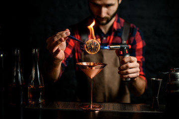 male barman holds tweezers with slice of citrus and sets fire to it
