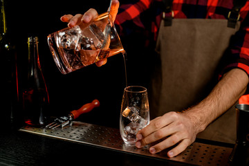 young male barman carefully pours cocktail from mixing cup into transparent glass.