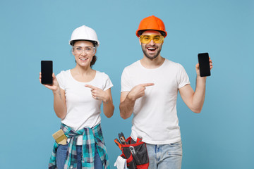 Funny couple woman man in hardhat glasses point index finger on mobile phone with blank empty screen isolated on blue background. Instrument accessories renovation apartment room. Repair home concept.