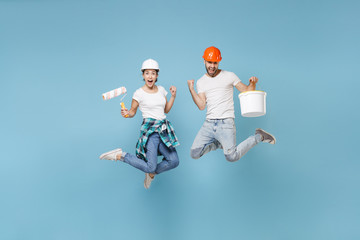 Couple woman man in protective helmet hardhat jump with paint bucket, roller isolated on blue background. Instruments accessories renovation apartment room. Repair home concept. Doing winner gesture.