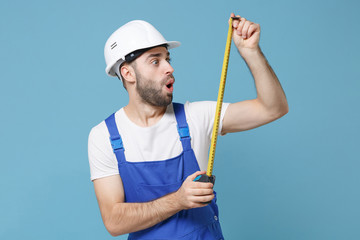 Shocked young man in coveralls protective helmet hardhat hold measure tape isolated on blue wall background studio portrait. Instruments accessories for renovation apartment room. Repair home concept.