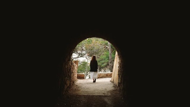 Girl in a white striped dress and grey jacket walks along the coastal path and passes through a stone tunnel. Long hair woman walks around the beautiful countryside. Golden light in idyllic landscape.