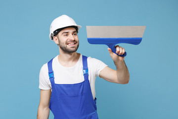 Smiling young man in coveralls protective helmet hardhat hold putty knife isolated on blue wall...