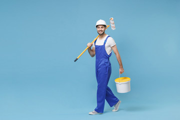 Smiling young man in coveralls protective helmet hardhat hold paint roller, bucket isolated on pastel blue wall background. Instruments accessories for renovation apartment room. Repair home concept.