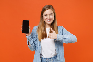 Naklejka premium Smiling young woman girl in casual denim clothes posing isolated on orange background studio. People lifestyle concept. Mock up copy space. Hold mobile phone with blank empty screen, showing thumb up.