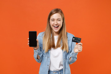 Laughing young woman girl in denim clothes isolated on orange background. People lifestyle concept. Mock up copy space. Hold mobile phone with blank empty screen, credit bank card keeping eyes closed.