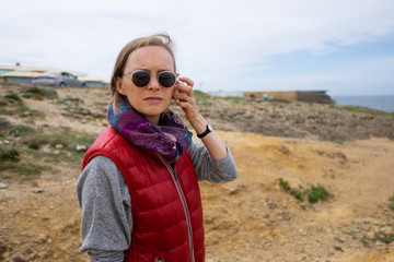 Frowning young woman wearing sunglasses and warm vest, walking on beach, looking at camera. People outdoors concept