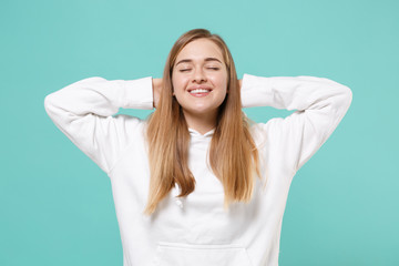 Smiling young woman girl in casual white hoodie posing isolated on blue turquoise background studio portrait. People emotions lifestyle concept. Mock up copy space. Sleeping with hands behind head.