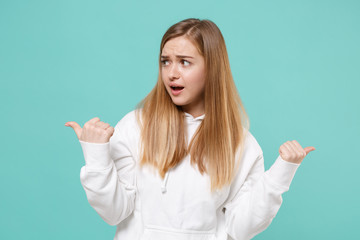 Perplexed young woman girl in casual white hoodie posing isolated on blue turquoise background studio portrait. People sincere emotions lifestyle concept. Mock up copy space. Pointing thumbs aside.