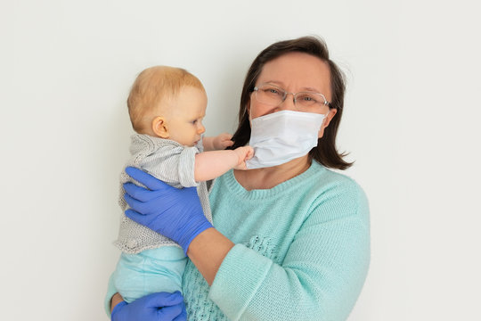 Babysitter Wearing Medical Mask And Gloves Holding Baby In Arms. Mature Woman With Little Child Posing Isolated Against White Background. Quarantine Or Public Health Concept