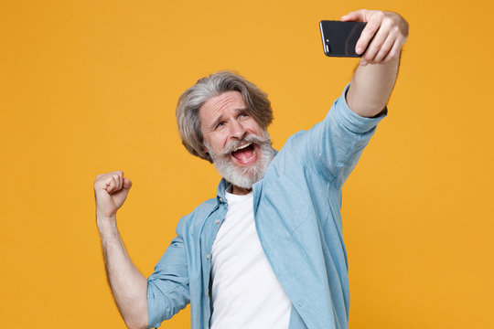 Happy elderly gray-haired mustache bearded man in casual blue shirt isolated on yellow background. People lifestyle concept. Mock up copy space. Doing selfie shot on mobile phone doing winner gesture.