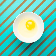 Raw egg in bowl on blue background with brown strips. Healthy food and cooking at home concept. Square format for Instagram.