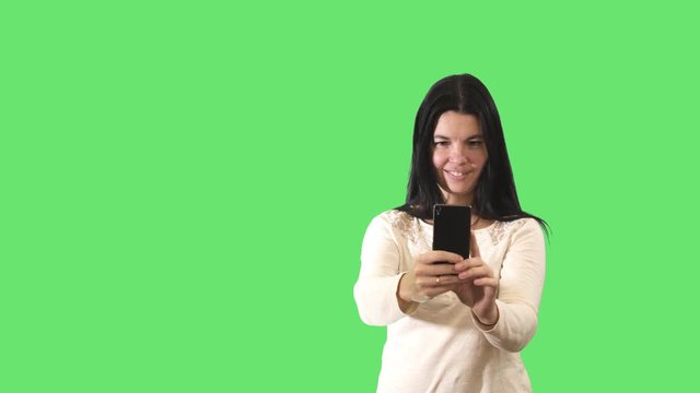 Smiling black-haired Caucasian woman taking photos with smartphone at green background. Portrait of joyful young lady photographing something. Leisure, tourism, chromakey, green screen.