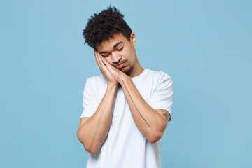 Tired young african american guy in casual white t-shirt posing isolated on pastel blue background studio portrait. People lifestyle concept. Mock up copy space. Sleep with folded hands under cheek.
