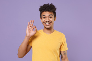 Smiling young african american guy in casual yellow t-shirt posing isolated on pastel violet background studio portrait. People emotions, lifestyle concept. Mock up copy space. Showing OK gesture.