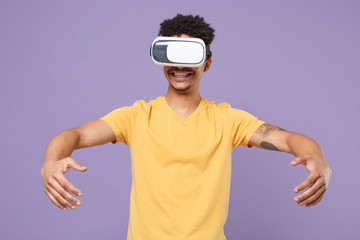 Smiling young african american guy in casual yellow t-shirt posing isolated on violet background. People lifestyle concept. Mock up copy space. Watching in headset spreading hands like hold something.