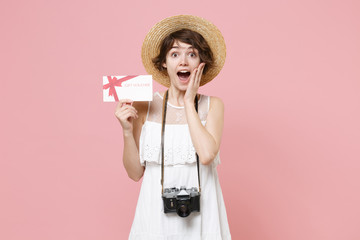 Excited young tourist girl in dress hat with photo camera isolated on pink background. Female traveling to travel weekends getaway. Air flight journey concept. Hold gift certificate put hand on cheek.
