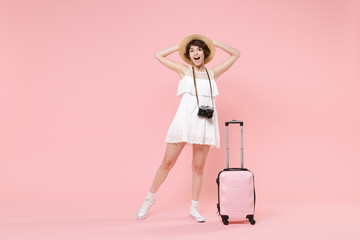 Excited young tourist girl in summer dress hat with photo camera suitcase isolated on pink background. Female traveling abroad to travel weekend getaway. Air flight journey concept. Put hands on head.