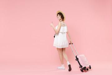 Back rear view of smiling tourist woman in summer dress hat with suitcase isolated on pink background. Female traveling abroad to travel weekends getaway. Air flight journey concept. Showing thumb up.
