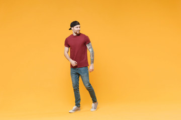 Fototapeta premium Cheerful young tattooed man guy in casual t-shirt black cap posing isolated on yellow wall background studio portrait. People sincere emotions lifestyle concept. Mock up copy space. Looking aside.