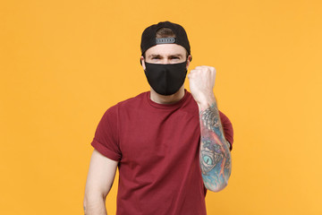Displeased young tattooed man guy in casual t-shirt cap black face mask posing isolated on yellow background studio portrait. People emotions lifestyle concept. Mock up copy space. Clenching fist.