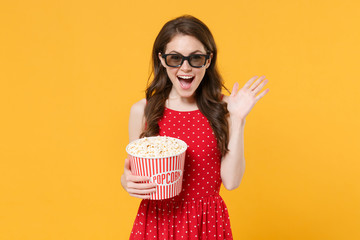 Excited brunette woman girl in red summer dress, 3d glasses isolated on yellow background. People lifestyle concept. Mock up copy space. Watching movie film, hold bucket of popcorn, spreading hands.