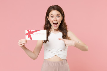 Surprised young brunette woman girl in light casual clothes posing isolated on pastel pink background studio. People lifestyle concept. Mock up copy space. Pointing index finger on gift certificate.